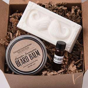 Square kraft brown box filled with brown paper shred and metal tin of beard balm, sample amber bottle of beard oil, bar of white goat milk soap with mustache design